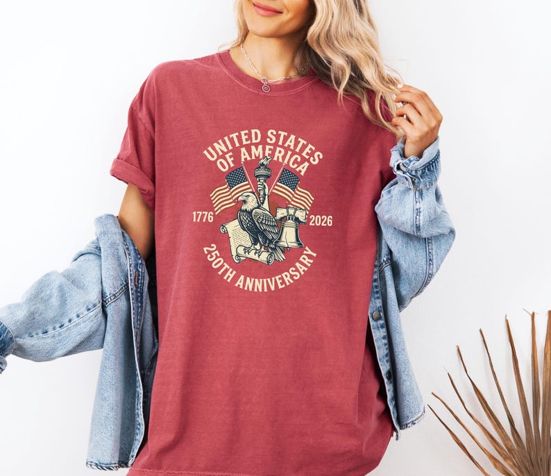 May include: A red t-shirt with a graphic commemorating the 250th anniversary of the United States of America. The design includes an eagle, flags, and the text "United States of America 250th Anniversary 1776-2026."