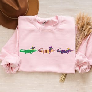 K&ouml;nnte beinhalten: Hellrosa Sweatshirt mit drei Cartoon-Alligatoren. Jeder Alligator ist mit Mardi Gras-Accessoires verziert: einer Krone, einem Hut und einer Krone. Das Sweatshirt hat lange &Auml;rmel und einen Rundhalsausschnitt.
