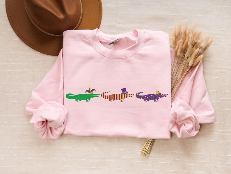 K&ouml;nnte beinhalten: Hellrosa Sweatshirt mit drei Cartoon-Alligatoren. Jeder Alligator ist mit Mardi Gras-Accessoires verziert: einer Krone, einem Hut und einer Krone. Das Sweatshirt hat lange &Auml;rmel und einen Rundhalsausschnitt.