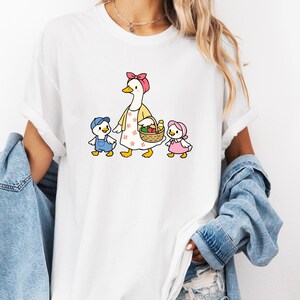 May include: White t-shirt with a cartoon illustration of a goose in a pink bow and yellow apron, carrying a fruit basket, and two ducklings in blue overalls and a pink bonnet. The design is centered on the shirt.