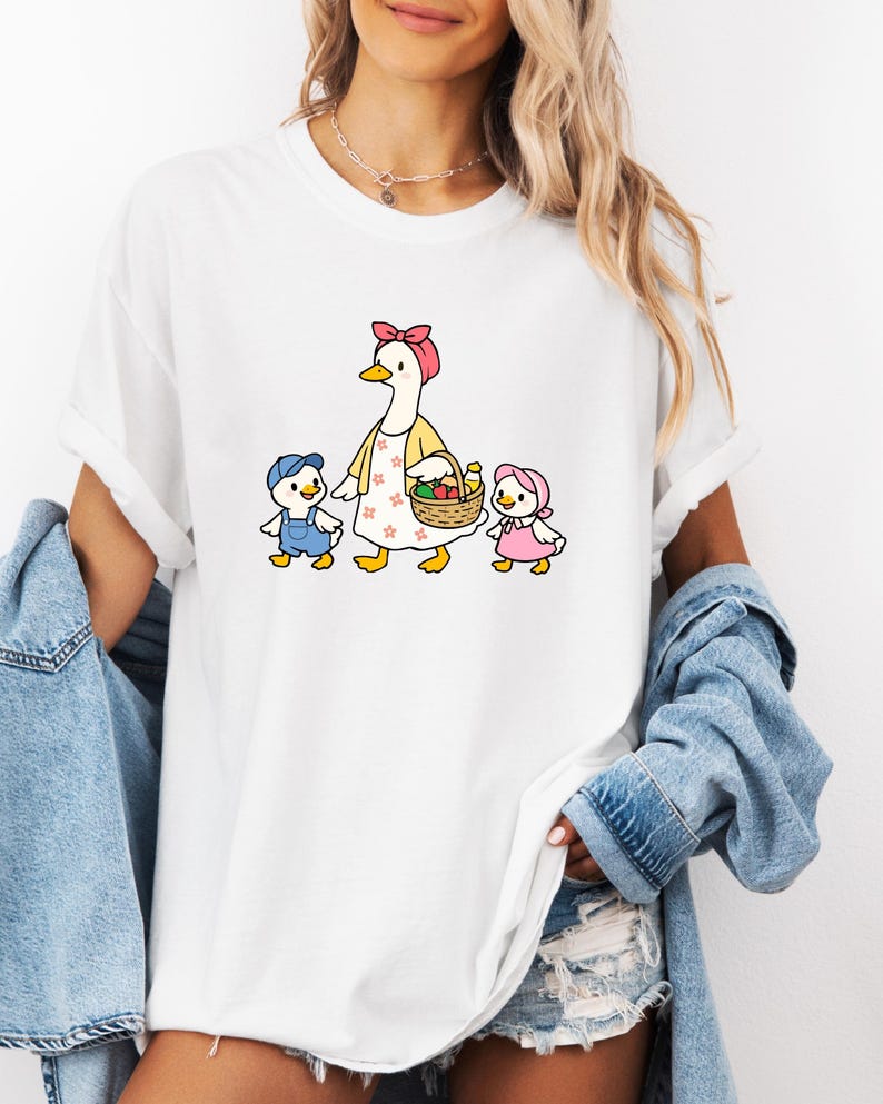 May include: White t-shirt with a cartoon illustration of a goose in a pink bow and yellow apron, carrying a fruit basket, and two ducklings in blue overalls and a pink bonnet. The design is centered on the shirt.