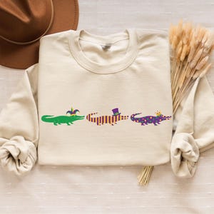 K&ouml;nnte beinhalten: Cremefarbenes Sweatshirt mit drei Cartoon-Alligatoren. Jeder Alligator ist mit Mardi-Gras-Accessoires verziert: einer Narrenkappe, einem Zylinder und einer Krone. Das Design ist in Gr&uuml;n, Lila und Gold gehalten.