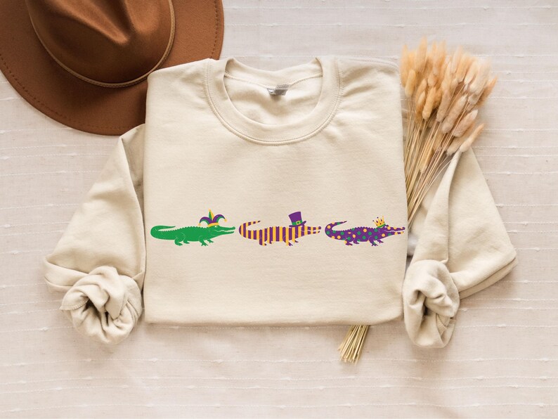 K&ouml;nnte beinhalten: Cremefarbenes Sweatshirt mit drei Cartoon-Alligatoren. Jeder Alligator ist mit Mardi-Gras-Accessoires verziert: einer Narrenkappe, einem Zylinder und einer Krone. Das Design ist in Gr&uuml;n, Lila und Gold gehalten.