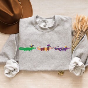 K&ouml;nnte beinhalten: Ein hellgraues Sweatshirt mit drei Cartoon-Alligatoren in Gr&uuml;n, Orange und Lila, die jeweils einen festlichen Hut tragen. Ein brauner Hut und Trockenblumen befinden sich im Hintergrund.