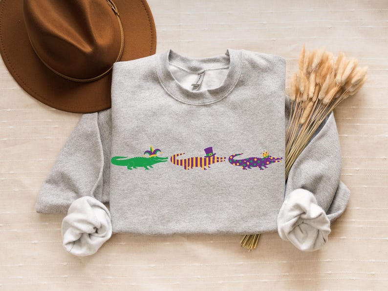 K&ouml;nnte beinhalten: Ein hellgraues Sweatshirt mit drei Cartoon-Alligatoren in Gr&uuml;n, Orange und Lila, die jeweils einen festlichen Hut tragen. Ein brauner Hut und Trockenblumen befinden sich im Hintergrund.