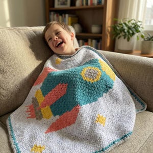 May include: A crocheted blanket featuring a teal rocket ship design with yellow, coral, and gray accents, set against a white background with yellow star details. The blanket has a teal border and is draped over a person.