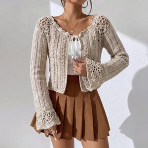 Puede incluir: Un cárdigan de crochet beige con mangas largas y escote festoneado. El cárdigan presenta un cierre de lazo y mangas acampanadas con un patrón floral de crochet. El conjunto incluye una falda plisada marrón.
