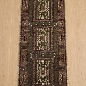 Puede incluir: Una alfombra rectangular tipo pasillero con un panel central verde oscuro que presenta tres motivos geométricos repetitivos en marrón y blanco. El borde es de color marrón oscuro con símbolos blancos y los extremos tienen flecos blancos.