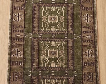 Puede incluir: Una alfombra rectangular tipo pasillero con un campo central verde oliva oscuro que presenta tres motivos geométricos cuadrados repetidos en marrón y crema. El borde es de color marrón oscuro con símbolos geométricos blancos y flecos.