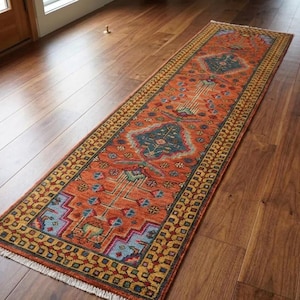 Può includere: Un lungo tappeto passatoia rettangolare caratterizzato da uno sfondo arancione vivace con intricati motivi geometrici e floreali nelle tonalità del blu, giallo e rosso. Il tappeto presenta un bordo decorativo e corte frange bianche alle estremità.