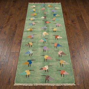 May include: A rectangular runner rug featuring a light green background decorated with a repeating pattern of stylized birds in various colors including blue, orange, yellow, and purple. The rug has a fringed edge on the top and bottom ends.