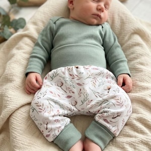 Pode incluir: Um bebé a dormir com um body verde sálvia de manga comprida e calças estampadas. As calças apresentam um fundo branco com um desenho floral em tons de castanho e verde. O bebé está deitado numa manta creme.