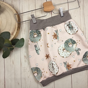 Può includere: Gonna rosa chiaro con fascia e orlo grigi, con un motivo di ciambelle, coniglietti, cuori e elementi floreali. La gonna è appesa a una gruccia di legno.
