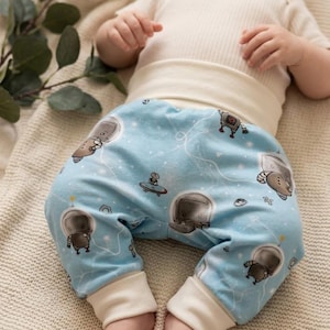 Può includere: Bambino con un body bianco e pantaloni azzurri con un motivo a tema spaziale. I pantaloni presentano astronauti dei cartoni animati, pianeti e stelle. Il bambino è sdraiato su una coperta color crema.