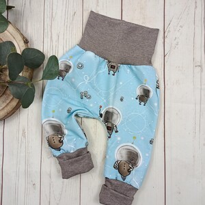 Può includere: Pantaloni da bambino con sfondo azzurro chiaro e motivo ripetuto di astronauti dei cartoni animati. I pantaloni hanno vita e polsini grigi. Il design include elementi a tema spaziale.
