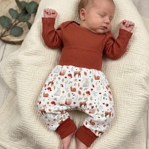 Pode incluir: Um bebé a dormir com um body de manga comprida canelado cor ferrugem e calças estampadas. As calças apresentam um fundo branco com criaturas da floresta, incluindo veados, corujas e coelhos. O bebé está deitado numa manta creme.