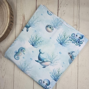 Puede incluir: Una manta de bebé plegada, de color azul claro, con ilustraciones marinas. El diseño incluye un caballito de mar, una ballena, un pulpo, una tortuga marina y un pez globo, con coral azul y burbujas. El fondo tiene un efecto acuarela azul claro.