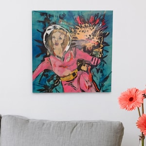May include: A colorful pop art painting of a woman in a pink space suit against a blue and orange background. The artwork hangs above a gray sofa, with a small white side table holding a vase of orange flowers.
