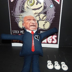 May include: A blue voodoo doll with a white face and a red tie. The doll has a black heart on its chest and is holding a stick with a blue ball on the end. The doll is sitting on a white surface with three white skull pins.