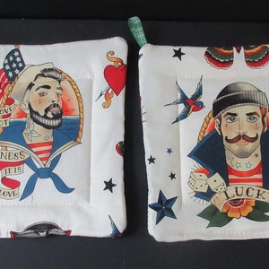 May include: Two white cotton pot holders with a nautical theme. Each pot holder features a sailor with a tattoo of a heart, an anchor, a swallow, dice, and a banner that says "Lucky".