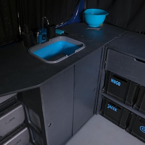 May include: A dark gray camper kitchen setup with a built-in sink, cabinets, and storage. The sink has a blue interior, and there's a blue bowl on the countertop. Storage bins are labeled with names. The overall design is modern and functional.