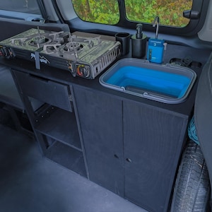 Op de afbeelding: Interieur van een camper met een ingebouwde keukenunit. De unit heeft een tweepits kookplaat, een opvouwbare blauwe wastafel en opbergkasten. Ontworpen voor kamperen, optimaliseert de unit ruimte en functionaliteit.
