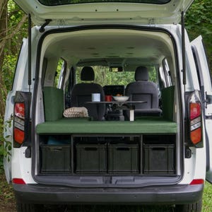 Può includere: L'interno di un furgone bianco convertito in camper, dotato di una panca imbottita verde, un tavolino nero con una ciotola e una tazza, e quattro cassette portaoggetti nere disposte in fila sotto la piattaforma per dormire.