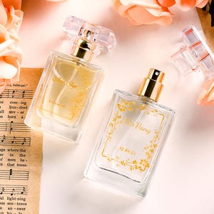 Puede incluir: Dos frascos de perfume de vidrio transparente con detalles dorados y diseños florales, uno con el texto "Annie Skye" y el otro "Miss Vang 12.19.23". Rosas melocotón y partituras musicales rodean los frascos.