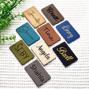 May include: A collection of rectangular name tags in various colors, including tan, brown, teal, olive green, cream, gray, and navy blue. Each tag features a different name in a contrasting color, such as black or gold, in a cursive font.