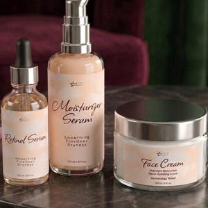 Peut inclure: Un ensemble de soins comprenant un flacon en verre transparent de sérum au rétinol avec compte-gouttes, un flacon pompe de sérum hydratant et un pot de crème pour le visage. Chaque contenant a une étiquette pêche avec du texte noir, indiquant 100 ml ou 120 ml.
