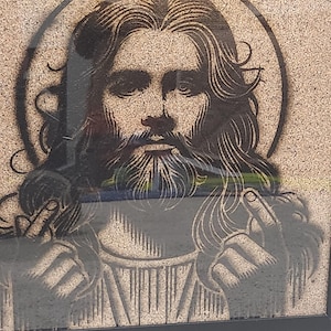 May include: Framed artwork featuring a black silhouette of a figure with long hair, a beard, and a halo. The figure's hands are raised, with the middle finger extended on each hand. The artwork is set against a textured, light brown background.