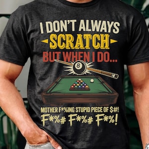 May include: A charcoal gray t-shirt featuring a graphic of a pool table with a rack of billiard balls and a cue stick. The text reads: I DON'T ALWAYS SCRATCH BUT WHEN I DO... MOTHER F*%ING STUPID PIECE OF $#! F*%# F*%# F*%#! in yellow and red.