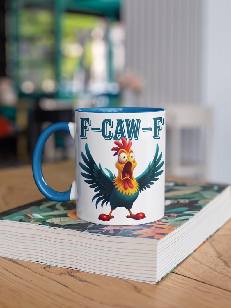 Pu&ograve; includere: Tazza in ceramica bianca con manico e interno blu. La tazza presenta un gallo dei cartoni animati con le ali aperte e la scritta "F-CAW-F" in blu. La tazza &egrave; appoggiata su un libro con una copertina colorata.