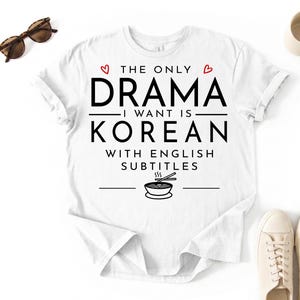 Könnte beinhalten: Weißes T-Shirt mit dem schwarzen Text "THE ONLY DRAMA I WANT IS KOREAN WITH ENGLISH SUBTITLES". Das Design beinhaltet zwei rote Herzen und eine Schüssel Nudeln mit Essstäbchen. Sonnenbrille und Schuhe sind sichtbar.