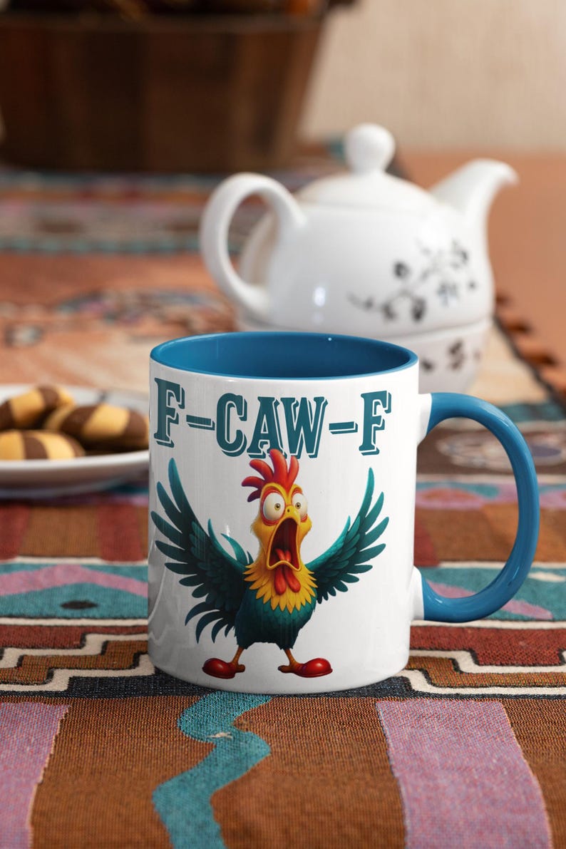 Pu&ograve; includere: Tazza in ceramica bianca con interno e manico blu, raffigurante un gallo dei cartoni animati con le ali aperte e la scritta "F-CAW-F". Una teiera e biscotti sono sullo sfondo. La tazza &egrave; su una tovaglia fantasia.