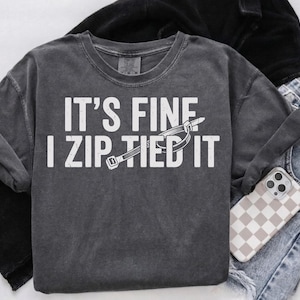 May include: Dark grey crew neck sweatshirt with the text "IT'S FINE I ZIP TIED IT" in white block letters and a zip tie graphic. The sweatshirt is folded with a pair of blue jeans and a phone case with a checkered pattern.