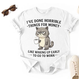 Puede incluir: Camiseta blanca con un gato de dibujos animados gruñón sosteniendo una taza de café. La camiseta tiene el texto "HE HECHO COSAS HORRIBLES POR DINERO COMO LEVANTARME TEMPRANO PARA IR A TRABAJAR". Gafas de sol y zapatos también son visibles.