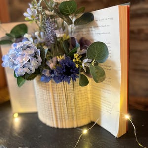 May include: A decorative floral display in a vase crafted from book pages. The arrangement includes blue and purple flowers with green leaves. An open book and fairy lights are also visible.