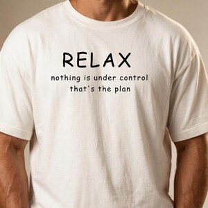 Könnte beinhalten: Ein cremefarbenes Kurzarm-T-Shirt mit dem schwarzen Text RELAX nothing is under control that's the plan auf der Brust. Das Oberteil hat einen Rundhalsausschnitt und eine lockere Passform, kombiniert mit einer olivgrünen Hose.