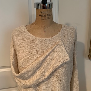 May include: A cream-colored knitted sweater with a loose fit and long sleeves. The sweater is made of a soft, textured yarn.