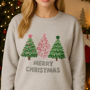 Pode incluir: Camisola cinza claro com três árvores de Natal verdes e rosas, com detalhes de estrelas e espirais. As palavras "MERRY CHRISTMAS" estão bordadas por baixo. A pessoa sorri em frente a uma árvore de Natal decorada.
