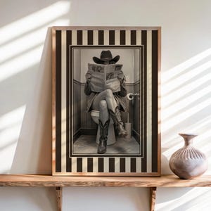 May include: A framed black and white wall art print featuring a person wearing a cowboy hat and boots, sitting on a toilet while reading a newspaper. The artwork is enclosed in a rectangular frame with a vertical black and white striped border.