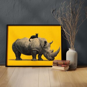 May include: A framed wall art print featuring a grey rhinoceros standing on a solid bright yellow background with a small black cat resting on its back. The artwork is displayed in a thin black frame, showcasing a high-contrast animal illustration.