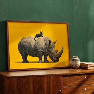 May include: A rectangular framed wall art print featuring a grey rhinoceros standing on a solid yellow background with a small black cat sitting on its back. The artwork is displayed in a simple brown wooden frame, showcasing a high-contrast design.