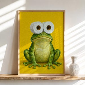 May include: A framed wall art print featuring a green frog with two white toilet paper rolls positioned as large eyes against a solid yellow background. The frog has a textured, painterly appearance with visible brushstrokes and a wide mouth.
