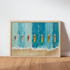 May include: A framed wall art print featuring an aerial view of seven figures on surfboards in various colors floating on textured blue ocean waves above a sandy beach. The surfboards are white, pink, and yellow, and the figures wear swimsuits.