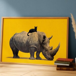 May include: A rectangular framed wall art print featuring a large grey rhinoceros standing on a solid bright yellow background with a small black cat sitting on its back. The frame is made of light-colored wood and is positioned on a wooden floor.