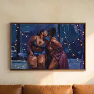 The image is a framed painting depicting two women in intimate poses, with one woman kissing the other. The painting is hung on a wall above a brown leather couch.