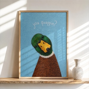 May include: A framed wall art print featuring a textured light blue background with the white text you quappin? above a mallard duck. The duck has a green head, a bright yellow beak, a white neck ring, and a brown body, all rendered in thick paint.