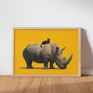 May include: A framed wall art print featuring a grey rhinoceros standing in profile against a solid mustard yellow background, with a small black cat resting on the back of the rhinoceros. The frame is made of light-colored natural wood.